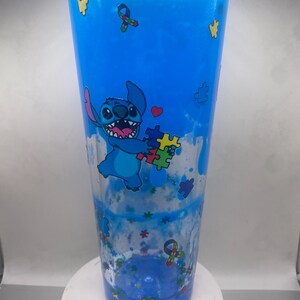 Puede incluir: Un vaso transparente lleno de l&iacute;quido azul y purpurina. El vaso presenta una imagen de dibujos animados de Stitch sosteniendo piezas de rompecabezas. El dise&ntilde;o incluye piezas de rompecabezas y cintas, con una combinaci&oacute;n de colores azul y transparente.