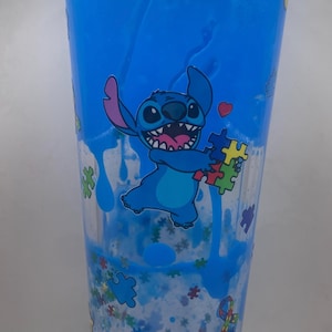 Puede incluir: Un vaso azul con el personaje Stitch sosteniendo piezas de rompecabezas, con un coraz&oacute;n. El vaso tiene un l&iacute;quido azul y purpurina en su interior. El dise&ntilde;o incluye gr&aacute;ficos de piezas de rompecabezas y cintas, y est&aacute; sobre una superficie blanca.