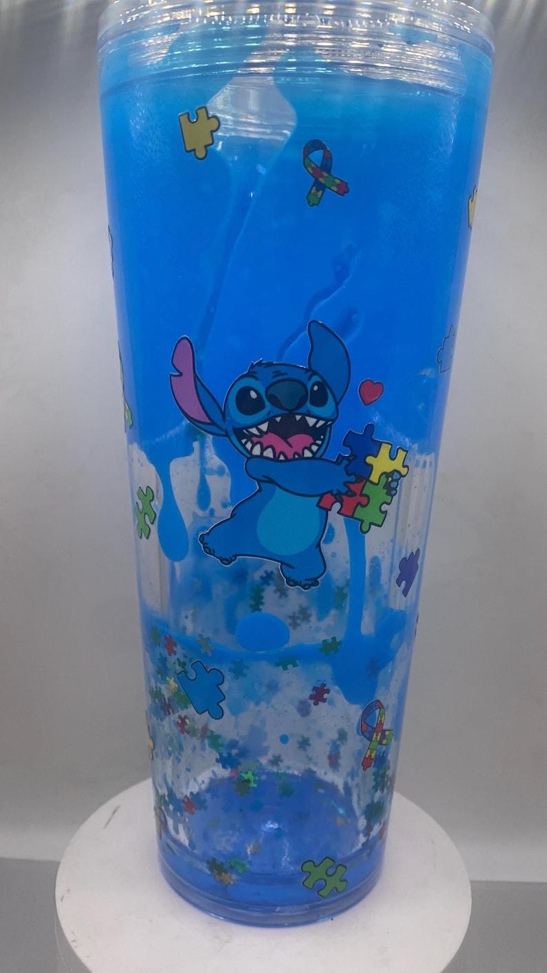 Puede incluir: Un vaso azul con el personaje Stitch sosteniendo piezas de rompecabezas, con un coraz&oacute;n. El vaso tiene un l&iacute;quido azul y purpurina en su interior. El dise&ntilde;o incluye gr&aacute;ficos de piezas de rompecabezas y cintas, y est&aacute; sobre una superficie blanca.