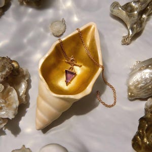 May include: A gold-colored necklace with a triangular purple gemstone pendant rests inside a seashell-shaped dish. The dish has a gold interior and a cream-colored exterior. Other decorative shells and objects surround the dish.