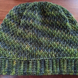 May include: A handmade beanie hat in shades of green, blue, and yellow. The hat features a ribbed brim and a textured, knitted body. The color palette gives the hat a natural, earthy look, suitable for cold weather. The hat is a cozy accessory.