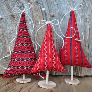 Könnte beinhalten: Drei dekorative Weihnachtsbaum-Dekorationen aus rotem Stoff mit verschiedenen Mustern, darunter Blumen- und geometrische Designs. Jeder Baum ist auf einem Holzsockel mit einem Holzdübel befestigt und mit einer weißen Schnurschleife gebunden. Die Bäume sind etwa 15 cm hoch.
