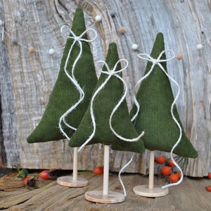 Könnte beinhalten: Drei handgefertigte grüne Stoff-Weihnachtsbäume mit weißen Schnurakzenten und Holzsockeln. Die Bäume sind auf einer rustikalen Holzoberfläche angeordnet, mit einer Kette aus Filzkugeln im Hintergrund. Die Bäume sind etwa 15 cm hoch.