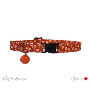 May include: Orange cat collar with a white floral pattern and a black buckle. The collar has a small orange bell attached.