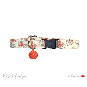 May include: A cream-colored cat collar with a floral pattern in shades of orange and gray. The collar has a black plastic buckle and a small orange bell.