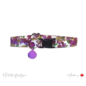 May include: A white cat collar with a purple floral pattern and a purple bell. The collar has a black buckle and is made in Canada.