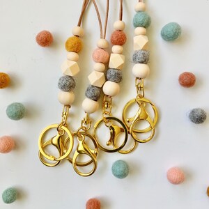 Może przedstawiać: Cztery brązowe sznurki ze skórzanymi paskami z drewnianymi i filcowymi koralikami w odcieniach szarego, różowego, pomarańczowego i zielonego. Każdy sznurek ma złoty metalowy brelok.