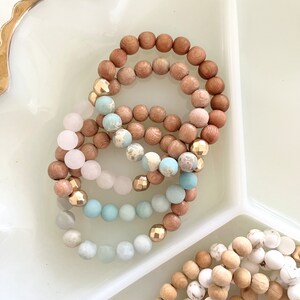 May include: A set of four beaded bracelets with wooden beads in natural and brown tones, and light blue and pink beads. The bracelets are stacked on top of each other on a white surface.