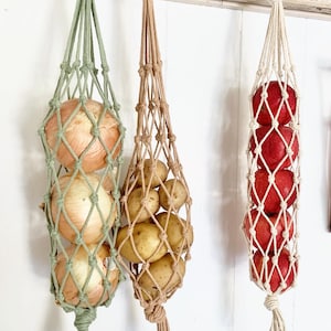 May include: Three hanging produce bags with different contents. A green bag holds three onions, a beige bag holds potatoes, and a white bag holds red apples.
