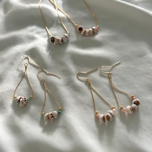 May include: Gold-toned necklaces and earrings featuring natural seashells. The necklaces have a delicate chain with a row of shells. The earrings have a teardrop design with shells and small beads. The jewelry is displayed on a white fabric.