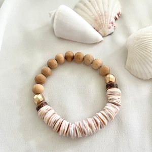 Può includere: Un braccialetto di legno e conchiglie con accenti dorati. Il braccialetto è su uno sfondo di tessuto bianco con conchiglie.