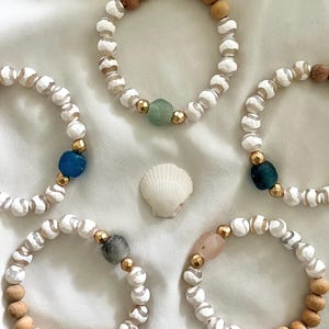 May include: Five bracelets with white and brown beads, each with a different colored gemstone accent. The bracelets are arranged on a white fabric background.