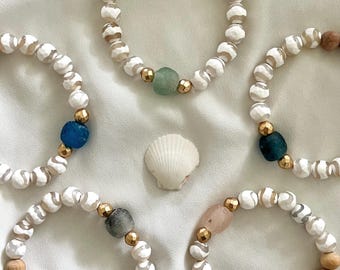 Può includere: Cinque braccialetti con perline bianche e marroni, ognuno con un accento di pietra preziosa di colore diverso. I braccialetti sono disposti su uno sfondo di tessuto bianco.