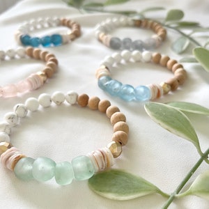 May include: A set of five bracelets featuring wooden beads and glass beads in shades of blue and green. The bracelets are displayed on a white fabric background with green foliage.
