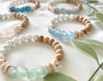 Può includere: Un set di cinque braccialetti con perline di legno e perline di vetro in tonalità di blu e verde. I braccialetti sono disposti su uno sfondo di tessuto bianco con foglie verdi.