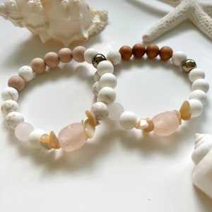 Könnte beinhalten: Zwei Armbänder mit Strand-Thema. Ein Armband hat weiße, rosa und beige Perlen, mit einem großen rosa Stein und Muschelakzenten. Das andere Armband hat weiße, braune und silberne Perlen. Muscheln und ein Seestern im Hintergrund.