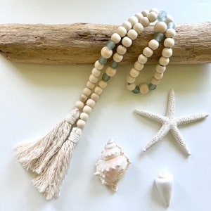 Puede incluir: Una guirnalda de cuentas de madera blanca con cuentas de vidrio azul claro y un borla blanca. La guirnalda se muestra en un trozo de madera a la deriva con una concha, una estrella de mar y una pequeña concha blanca.