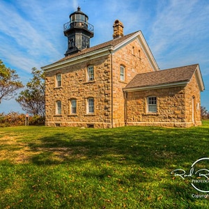 May include: A historic stone lighthouse with a black tower and a light on top. The building has multiple windows and a chimney, set against a blue sky with wispy clouds. The lighthouse sits on a grassy hill.