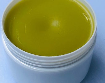 May include: Open white plastic jar filled with a smooth, light green substance. The jar is set against a light blue background. The product appears to be a cosmetic or skincare item.