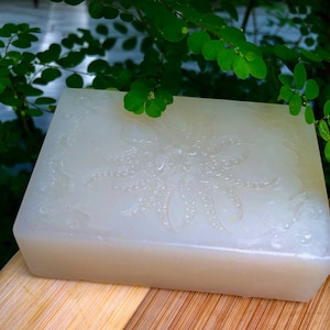 May include: A white bar of soap with a floral design embossed on the top. The soap is sitting on a wooden surface.
