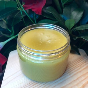 Puede incluir: Un tarro de vidrio transparente lleno de un bálsamo o ungüento amarillo. El tarro está sobre una superficie de madera clara.