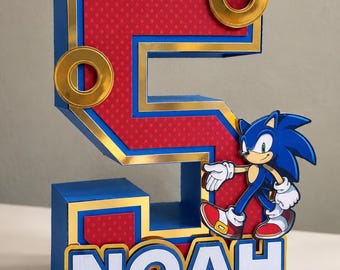 May include: A three-dimensional number five decoration featuring a red dotted pattern, blue borders, and gold metallic accents. It includes two gold rings and a cutout of the character Sonic the Hedgehog. The name NOAH is printed in white.