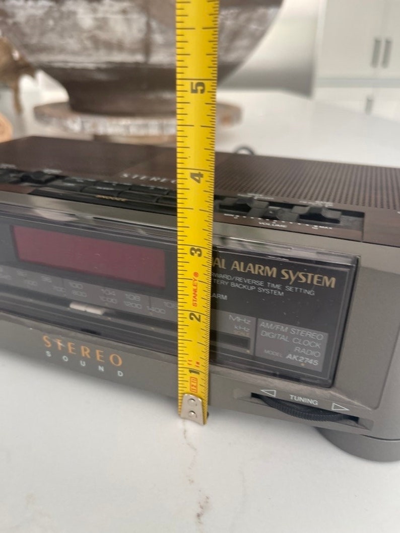 Pu&ograve; includere: Un radio digitale stereo AM/FM grigio e nero con un display digitale rosso. La radio ha un numero di modello AK2745 e dispone di una manopola di sintonizzazione e un sistema di allarme.
