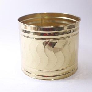 May include: A cylindrical, gold-colored metal container with a wavy pattern on the sides. The container has a shiny, reflective surface and is likely used for storage or decoration. The design includes horizontal bands near the top and bottom.