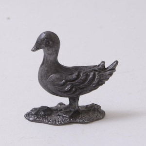 May include: A small, detailed metal duck figurine in a dark gray color. The duck is standing on a textured base, with its wings and feathers intricately sculpted. The figurine is a decorative item, likely for display.
