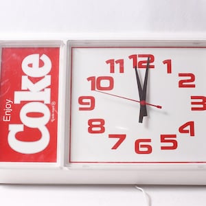 May include: White rectangular wall clock with a red and white Coca-Cola logo on the left and a clock face on the right. The clock face has red numbers and black hands, showing the time as nearly 12 o'clock.
