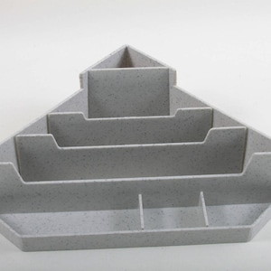 May include: A light gray desk organizer with multiple compartments. The organizer has a triangular shape with several tiered sections and small dividers, ideal for storing office supplies. The speckled texture adds visual interest.