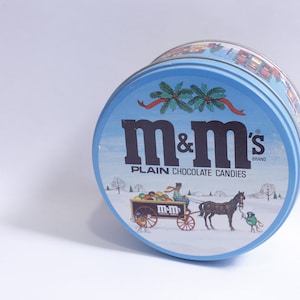 Może przedstawiać: Okrągłe, niebiesko-białe pudełko z zimową sceną i powozem ciągniętym przez konia. Pudełko zawiera napis "m&m's" czarnymi i białymi literami oraz "PLAIN CHOCOLATE CANDIES" czarnymi literami.