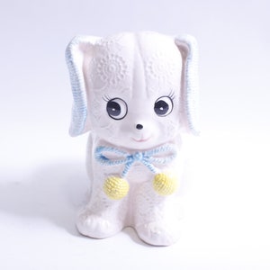 Può includere: Figurina di cucciolo di ceramica bianca con un fiocco blu e bianco e pompon gialli. Il cucciolo ha una texture a pizzo e grandi occhi neri.