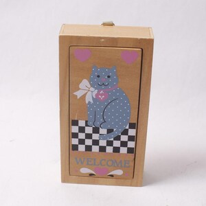 Könnte beinhalten: Eine rechteckige Holzschachtel mit Schiebedeckel, verziert mit einer blauen, gepunkteten Katze. Die Katze trägt eine weiße Schleife und einen rosa Herzanhänger. Darunter befinden sich ein schwarz-weißes Schachbrettmuster, das Wort WELCOME und rosa Herzen.