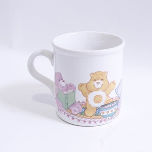 May include: White ceramic mug with a colorful illustration of three Care Bears. The Care Bears are sitting on a pink and white blanket with a bowl of cereal and a book. The text "Care Bears" is printed on the bottom of the mug.