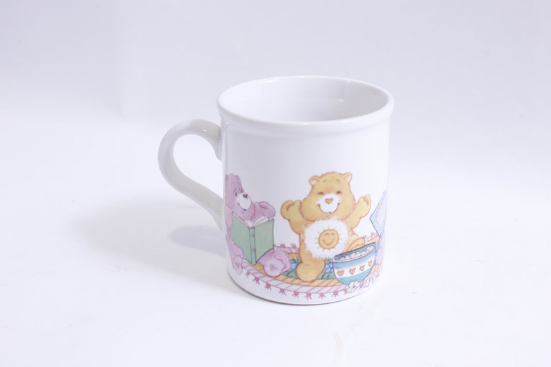 May include: White ceramic mug with a colorful illustration of three Care Bears. The Care Bears are sitting on a pink and white blanket with a bowl of cereal and a book. The text "Care Bears" is printed on the bottom of the mug.