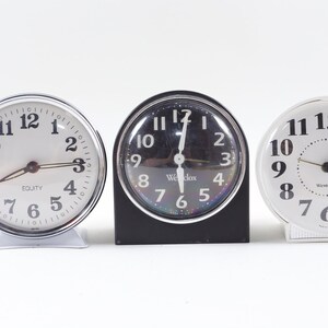 May include: Three vintage alarm clocks, one black, one white, and one silver, all with black numbers and hands. The clocks are all set to different times.