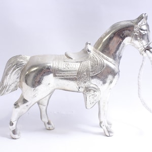 Może przedstawiać: Srebrna figurka konia z siodłem i wodzami. Koń stoi na białym tle.