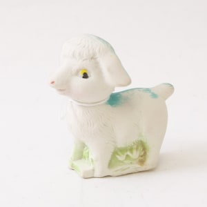 May include: A white ceramic lamb figurine with blue and green accents. The lamb has a yellow eye and is standing on a green base.