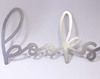 Puede incluir: Una pieza de arte de pared de metal plateado que deletrea la palabra "books" en una fuente cursiva. Las letras están conectadas y tienen un diseño moderno y minimalista. La obra de arte se muestra sobre un fondo blanco.