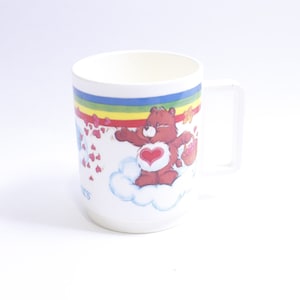 May include: White ceramic mug with a Care Bear design. The bear is brown with a red heart on its chest and is holding a red heart-shaped balloon. The mug has a rainbow and clouds around the bear.