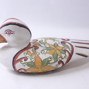 Puede incluir: Un pato de madera blanco con diseños florales pintados a mano en tonos rojos, naranjas, amarillos y verdes. El pato tiene un ojo negro y un pico rojo.