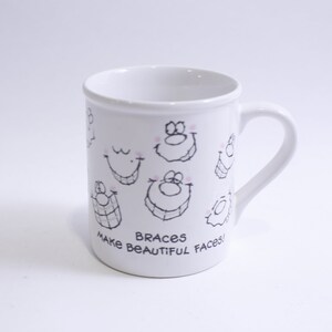 Puede incluir: Taza de cerámica blanca con asa, decorada con caras de dibujos animados y el texto "Braces make beautiful faces!" en negro. La taza presenta varias caras sonrientes con aparatos ortopédicos, lo que añade un toque lúdico al diseño.
