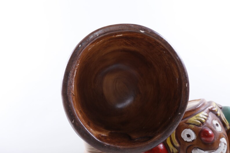 Op de afbeelding: Een bruin keramieken clownbeeldje met een grote, open mond trompet. De clown heeft een geschilderd gezicht met een rode neus, witte tanden en kleurrijk haar. De trompet is donkerbruin van kleur.