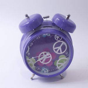 May include: Purple alarm clock with a peace sign and flower design. The clock has a white face with black numbers and hands.