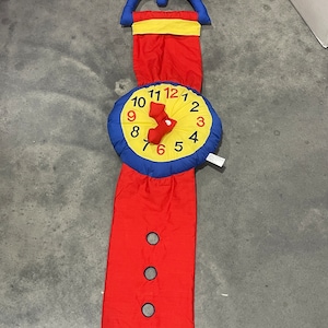 May include: A red, yellow and blue fabric clock with a hanging loop. The clock face has numbers 1-12 and a red hand.