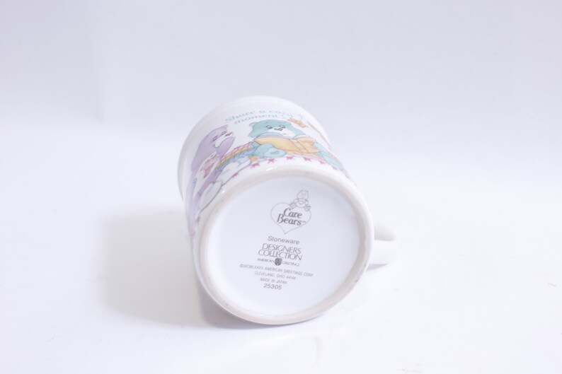 May include: White ceramic Care Bears mug with a colorful illustration of the Care Bears. The mug has a handle and the text "Stoneware Designers Collection" on the bottom.