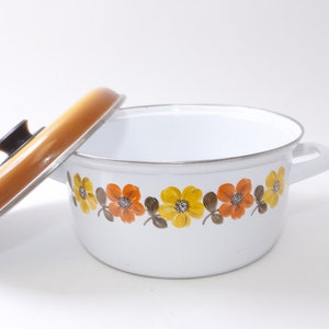 Könnte beinhalten: Ein weißer Emailletopf mit einem braunen und gelben Blumendesign am Rand. Der Topf hat zwei Griffe und einen passenden Deckel.