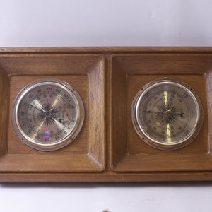 Puede incluir: Un barómetro enmarcado en madera con dos manómetros analógicos redondos. Los manómetros tienen biseles plateados y caras blancas con marcas negras. El manómetro izquierdo tiene un diseño de sol y luna y el manómetro derecho tiene un diseño de brújula.