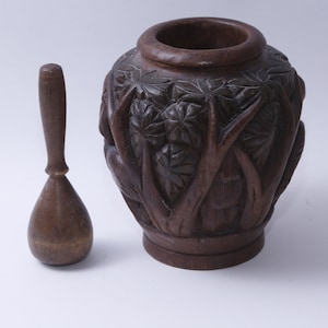 Könnte beinhalten: Ein dunkles braunes, aus Holz geschnitztes Mörser- und Stößel-Set mit aufwendigen Schnitzereien von Blättern und Ranken. Der Mörser ist vasenförmig und der Stößel hat eine abgerundete Form.